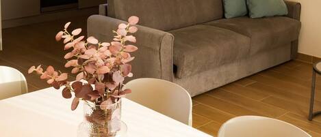 Una sala moderna con sofá, una mesa de comedor con sillas, un jarrón con flores, una zona de cocina con armarios y microondas, y una lámpara fijada a la pared.