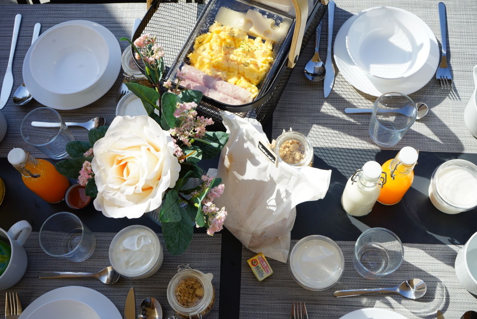 A breakfast table set with plates, cutlery, a glass of orange juice, a glass of milk, a jar of jam, a bowl of cereal, and a bouquet of flowers.