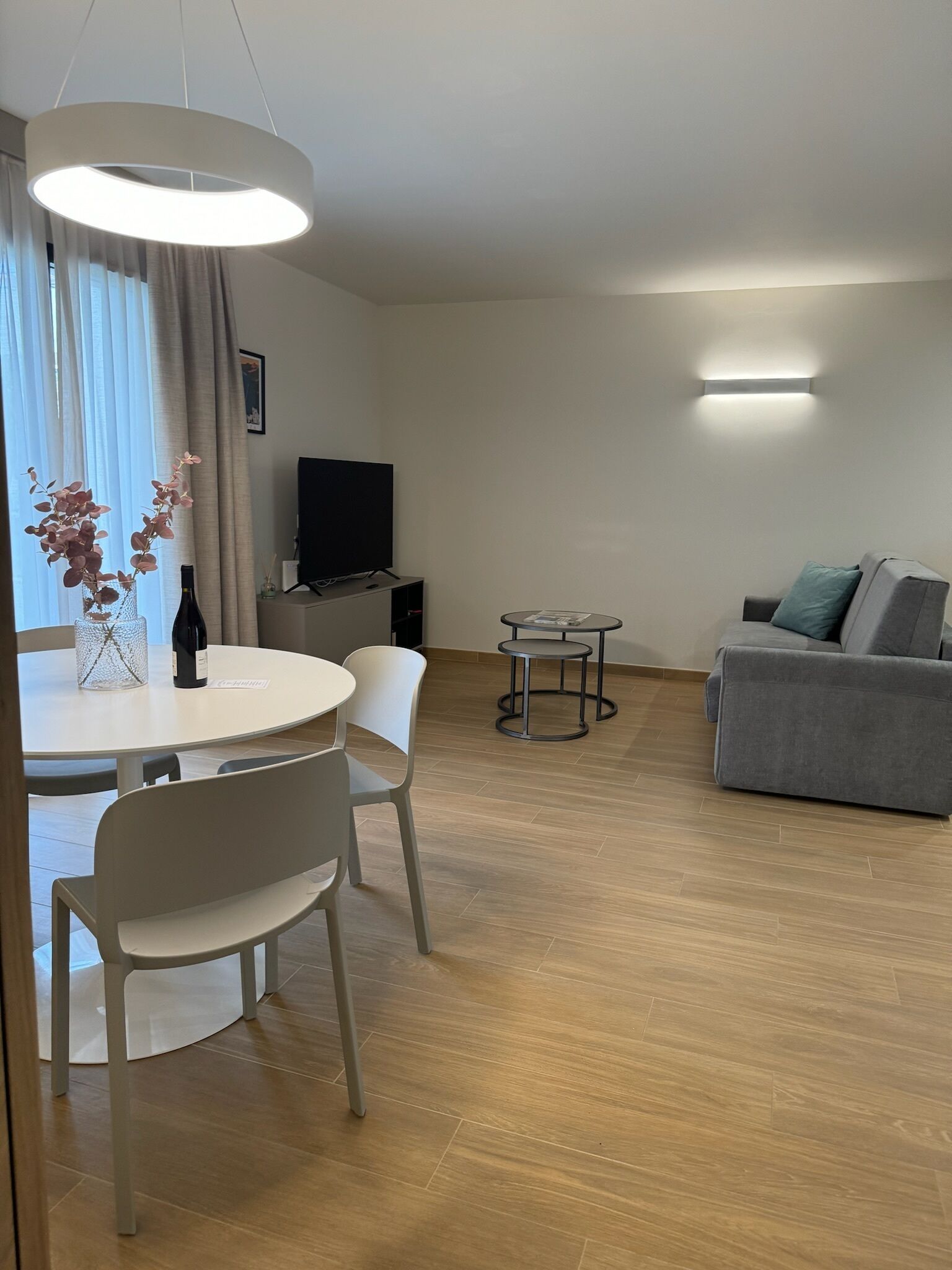 Un salon moderne avec un canapé gris, une table de salle à manger ronde entourée de chaises, un téléviseur à écran plat et un luminaire suspendu.