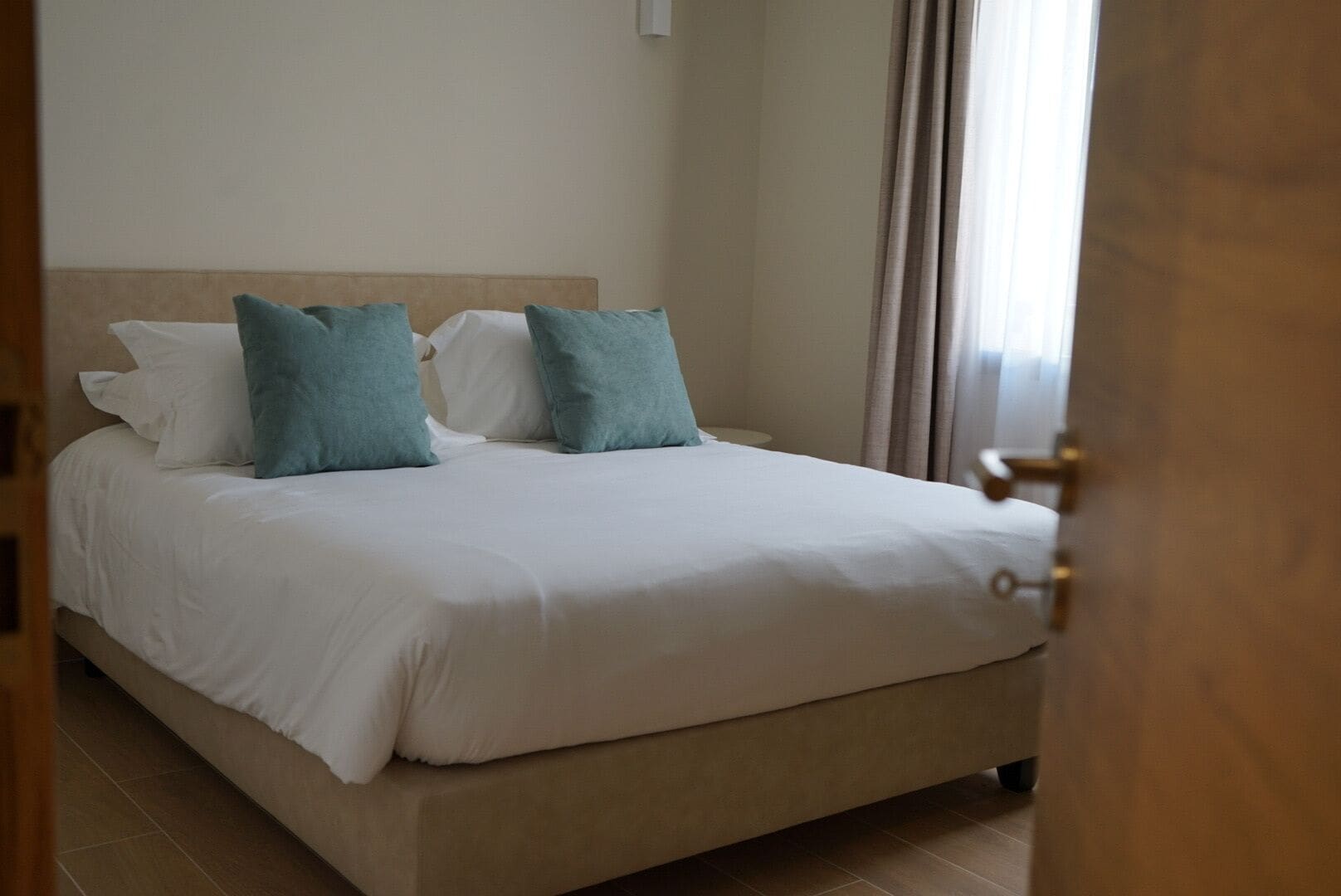 Un letto rifatto con cuscini bianchi e color acquamarina, una testiera beige e uno stipite in legno.