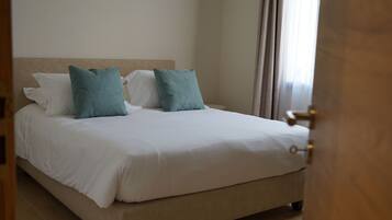 A neatly made bed with white and teal pillows, a beige headboard, and a wooden door frame.