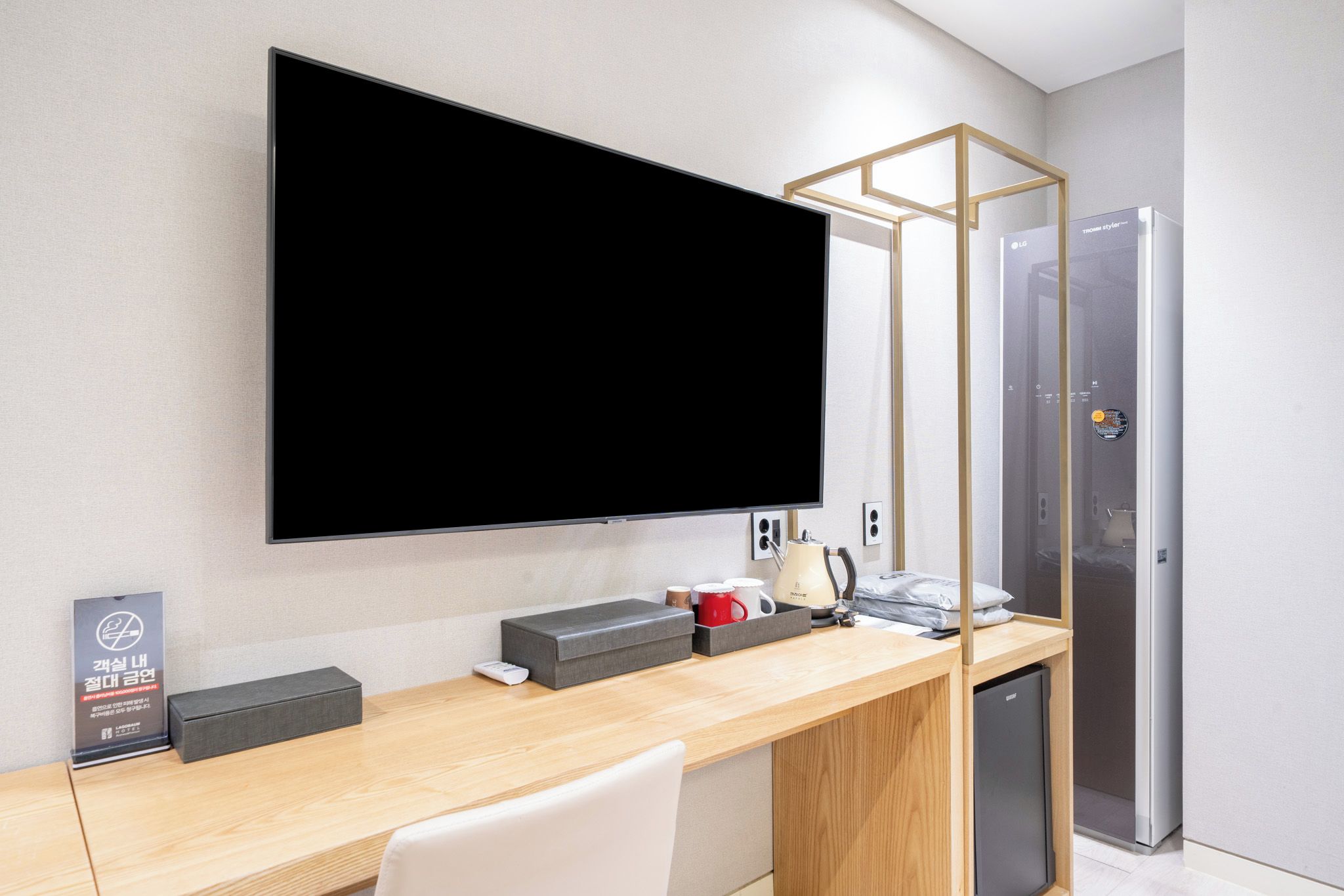 Una habitación de hotel moderna con un televisor de pantalla plana grande montado en la pared, un escritorio de madera con una silla, un refrigerador pequeño y una kitchenette compacta.