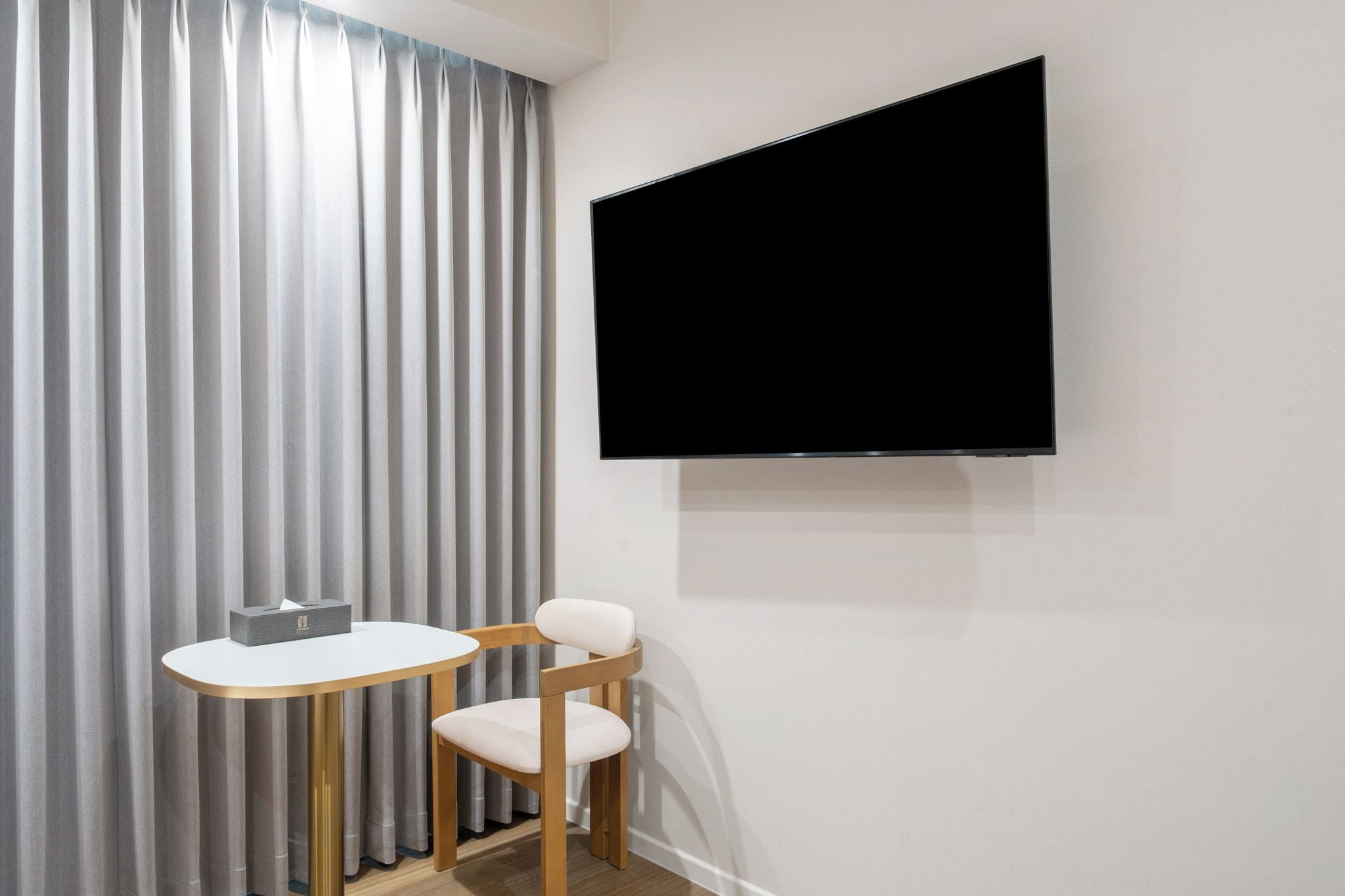 A room with a flat-screen TV mounted on the wall, a small round table with a chair, and a set of vertical blinds.