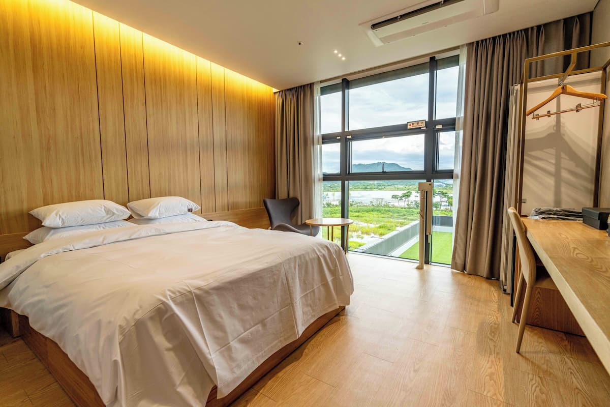 Habitación de hotel moderna con una cama grande, un escritorio y vistas a un paisaje verde a través de un ventanal amplio.