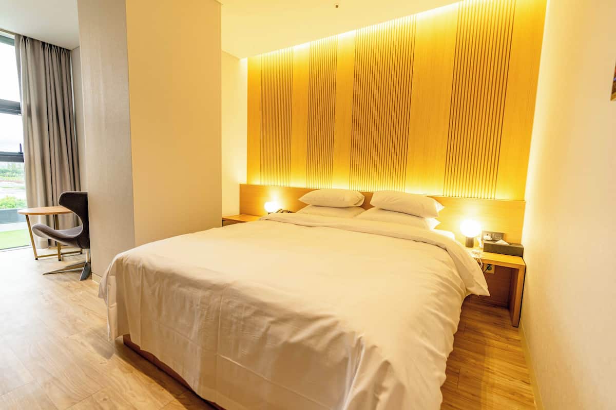 Un moderno cuarto de hotel con una cama grande, una mesita de noche con lámpara, una silla y vistas al exterior a través de una ventana con cortinas.