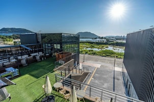 Una terraza en la azotea con mobiliario exterior, un área con paredes de vidrio y vistas a una masa de agua y colinas distantes.