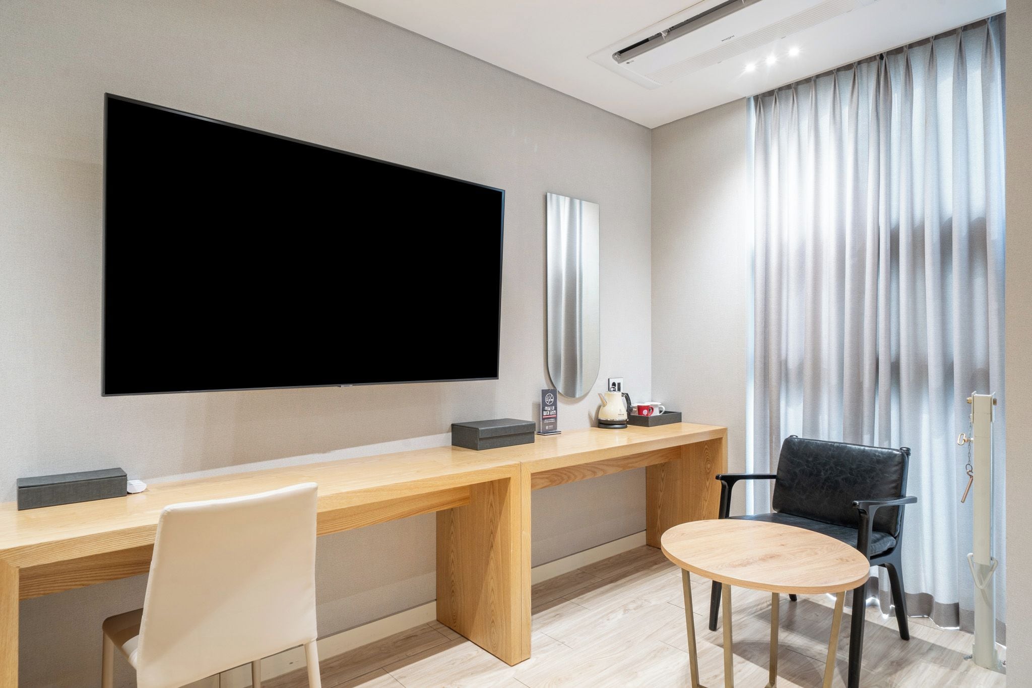 Ett hotellrum med en stor platt-TV som är monterad på väggen, ett skrivbord i trä med en stol, ett mindre runt bord och en svart läderstol.