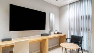 Ett hotellrum med en stor platt-TV som är monterad på väggen, ett skrivbord i trä med en stol, ett mindre runt bord och en svart läderstol.