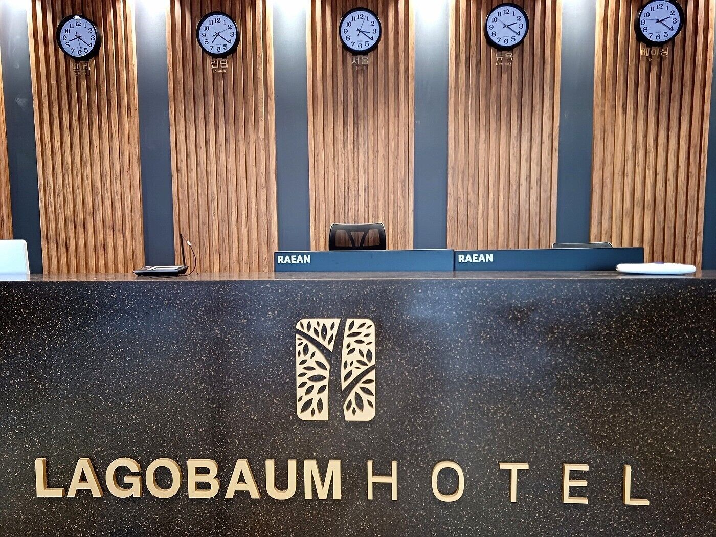 A reception desk with a wooden panel background and clocks displaying different time zones. The name "LAGOBAUM HOTEL" is prominently displayed on the desk.
