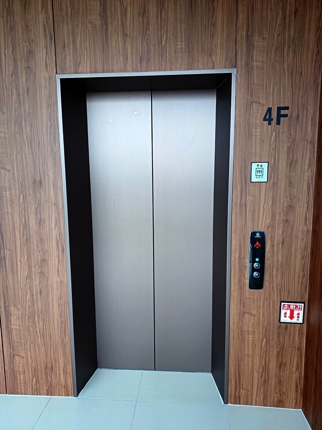 Un ascensor con interior metálico, un panel de madera a la derecha y un tablero de control con instrucciones de emergencia.