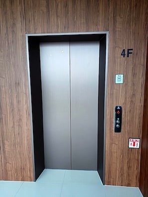 An elevator with a metallic interior, a wooden panel on the right, and a control panel with emergency instructions.