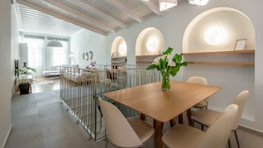 Un espacio comedor moderno con una mesa de madera, sillas color beige y un jarrón de flores. Hay lámparas del techo en arco y se ve otra habitación con un sofá y una planta.