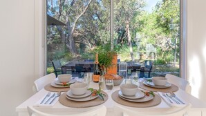Mesa de comedor para cuatro personas con platos, recipientes y cubiertos blancos, con vistas a una zona boscosa.