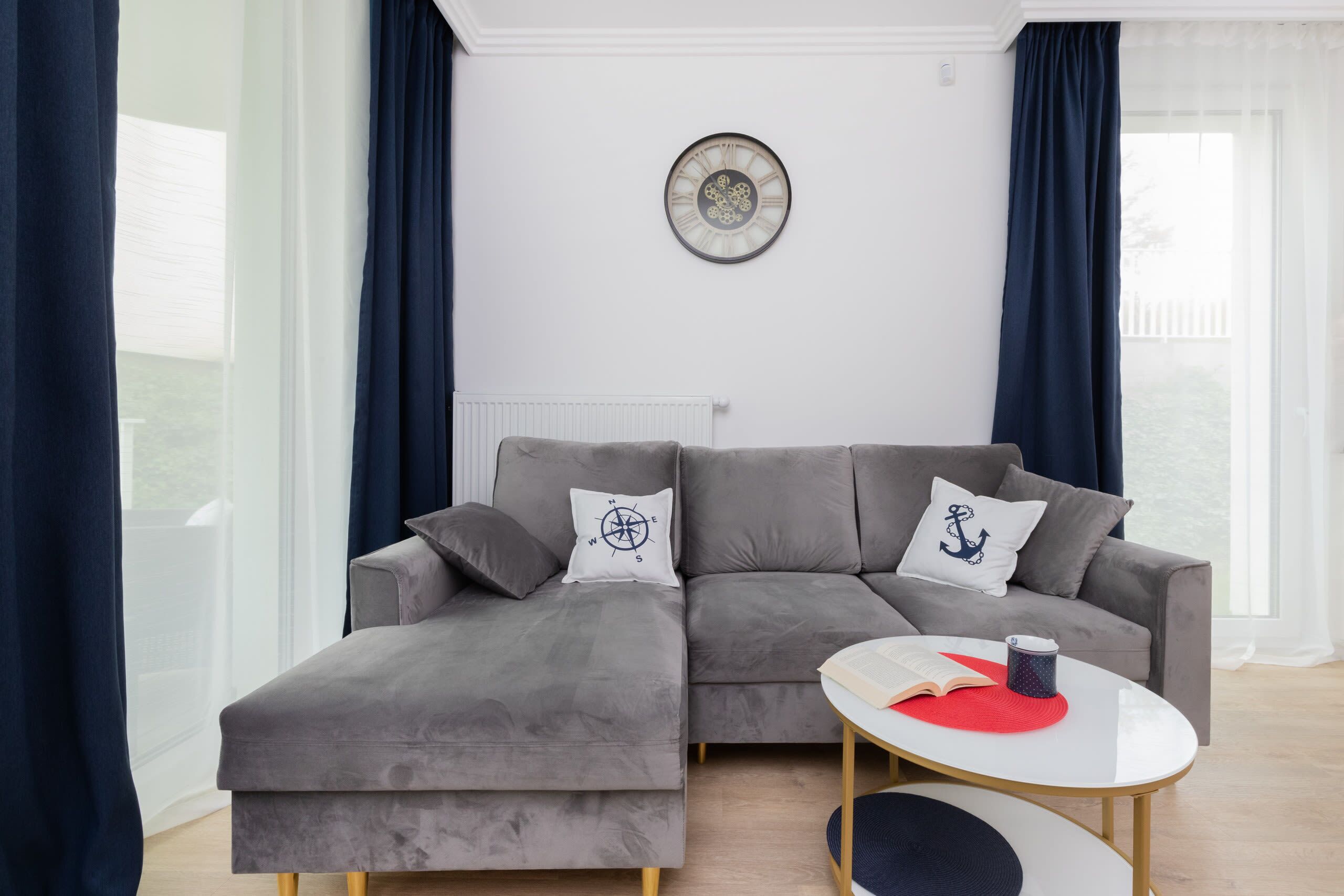 A modern living room with a grey sectional sofa, a round wall clock, and a small round table with a book and a cup.