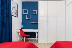 Une chambre avec un mur d’accent bleu, une armoire blanche, un bureau avec une chaise rouge et deux lits rouges.