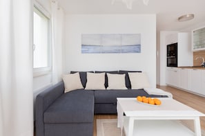 A modern living room with a grey sectional sofa, a white coffee table with oranges, a kitchen with white cabinets, and a painting of a seascape on the wall.