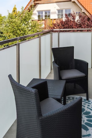 Ein Balkon mit zwei Korbstühlen und einem kleinen Tisch, einer weißen Wand und einem Gebäude mit Fenstern im Hintergrund.