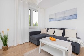 A modern living room with a grey sectional sofa, a white coffee table with oranges, a large window with white curtains, and a painting on the wall.