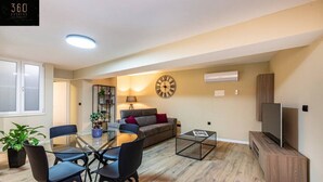 Ein modernes Wohnzimmer mit einem runden Glastisch, schwarzen Stühlen, einem grauen Sofa, einem Holztisch und einem Fernseher auf einem Holzschrank. Eine große Wanduhr ist ebenfalls zu sehen.