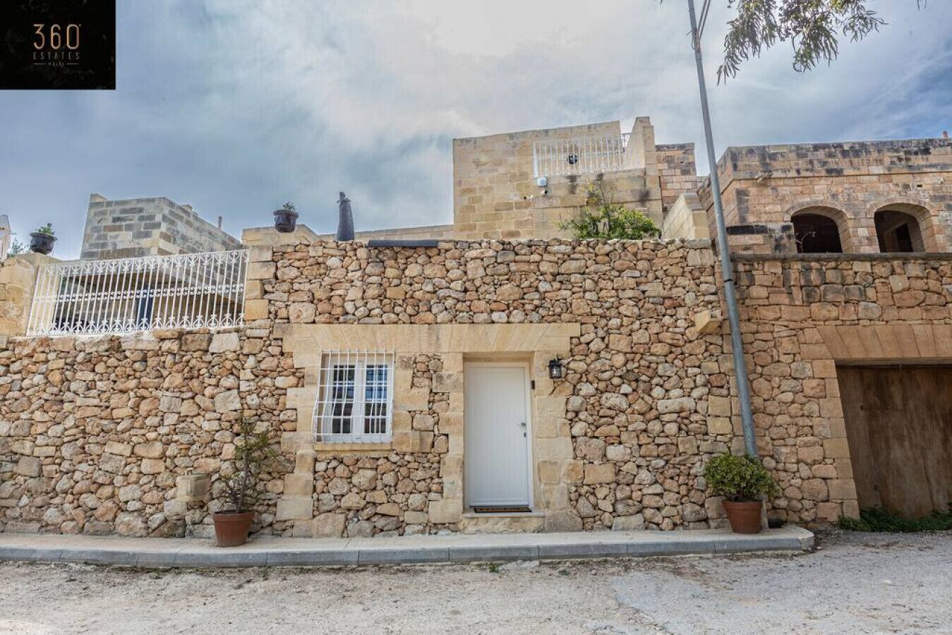 A stone building with a white door and window, potted plants, and a metal fence.