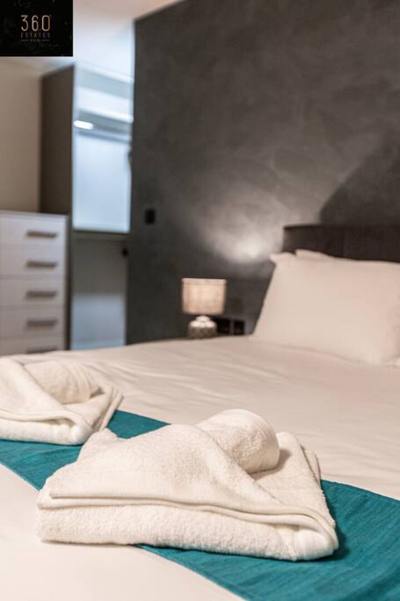 Una cama bien hecha con una colcha azul verdosa, una almohada blanca y una toalla blanca doblada.