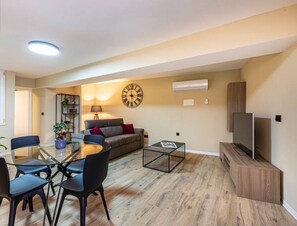 A modern living room with a round glass table, black chairs, a grey sofa, a wooden coffee table, a TV on a wooden stand, and a large wall clock.
