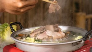 Una persona sosteniendo un palillo sobre una olla con sopa hirviendo, se aprecian ingredientes como carne y verduras.
