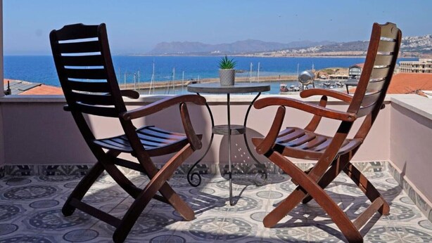 Deux chaises en bois et une petite table sur un balcon offrant une vue sur la mer et les montagnes lointaines.