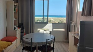 A room with a round table and chairs, a bookshelf, a bed, and a view of the beach. - Logement Front de mer à Port Leucate (Leucate)