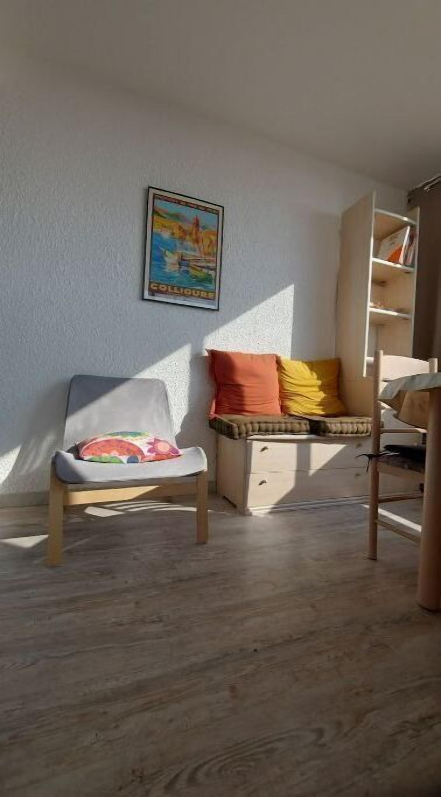 Une pièce chaleureuse avec un plancher en bois, un fauteuil confortable agrémenté d’un coussin coloré, un petit canapé avec un coussin à motifs, une bibliothèque et une affiche accrochée au mur.