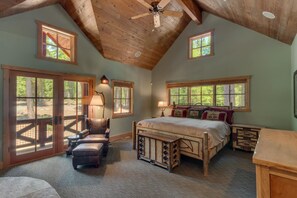 Une chambre spacieuse avec un grand lit, une tête de lit en bois et un banc en bois. Deux fenêtres laissent entrer la lumière naturelle, et un ventilateur de plafond est visible.
