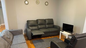 Ein Wohnzimmer mit einem grauen Sofa, einem schwarzen Sessel, einem grauen Teppich und einem Fernseher auf einem weißen Tisch.