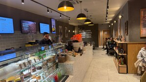 A modern coffee shop interior with a counter displaying pastries and beverages, a menu board, and a seating area. - Lavender Airport Hotel (Arnavutköy)