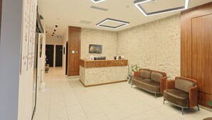 A reception area with a marble wall, a reception desk, two leather armchairs, and a ceiling with geometric lighting. - Lavender Airport Hotel (Arnavutköy)