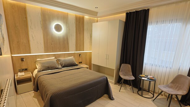 A modern bedroom with a large bed, bedside tables, a round mirror, a wardrobe, a small round table, two chairs, and a large window with curtains. - Lavender Airport Hotel (Arnavutköy)