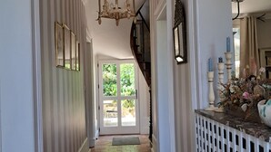 A hallway with a brick floor, white walls, and a chandelier. There is a door at the end of the hallway leading outside.