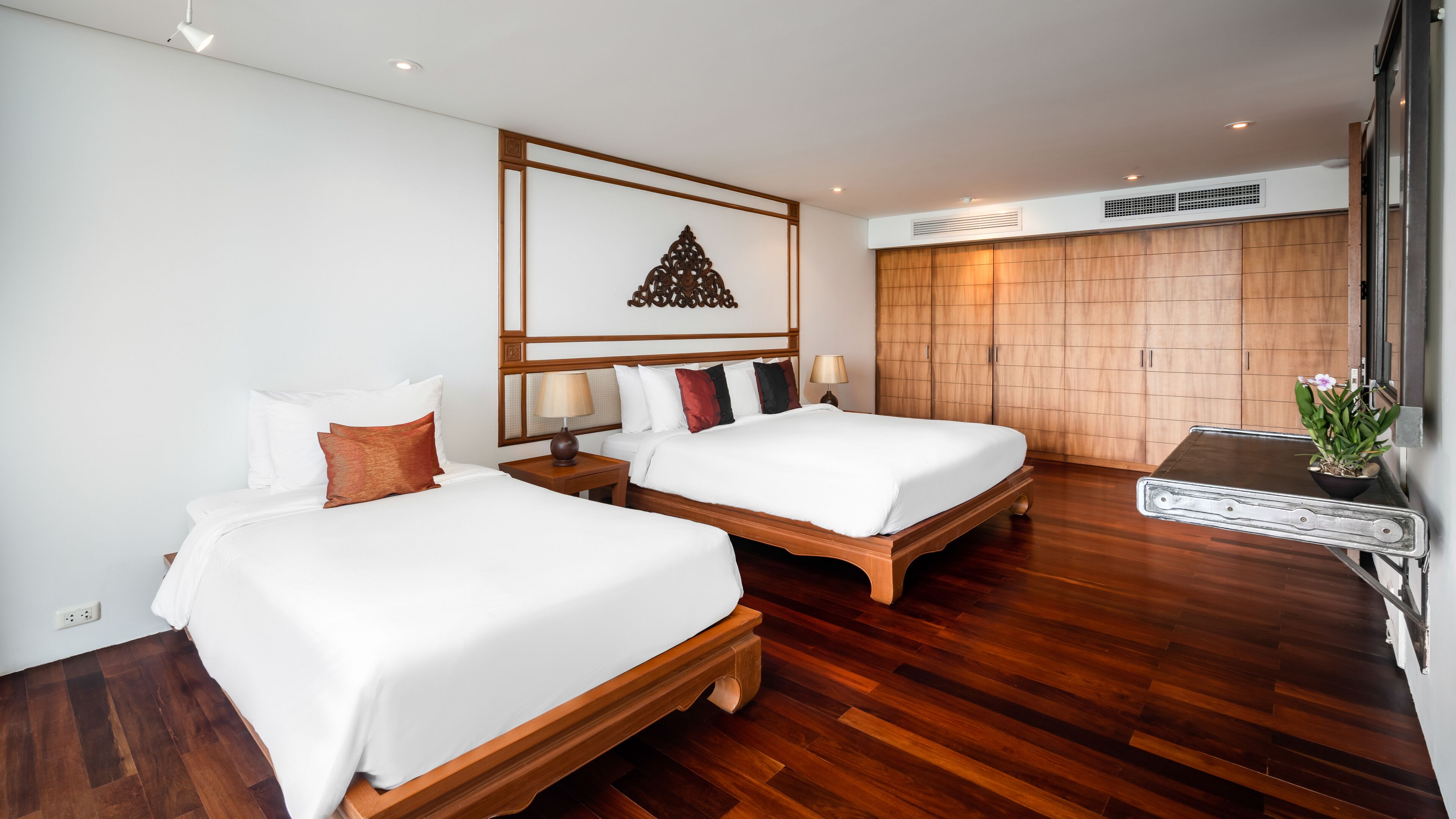 A hotel room with two beds, wooden flooring, a decorative wall panel, and a small table with a plant.