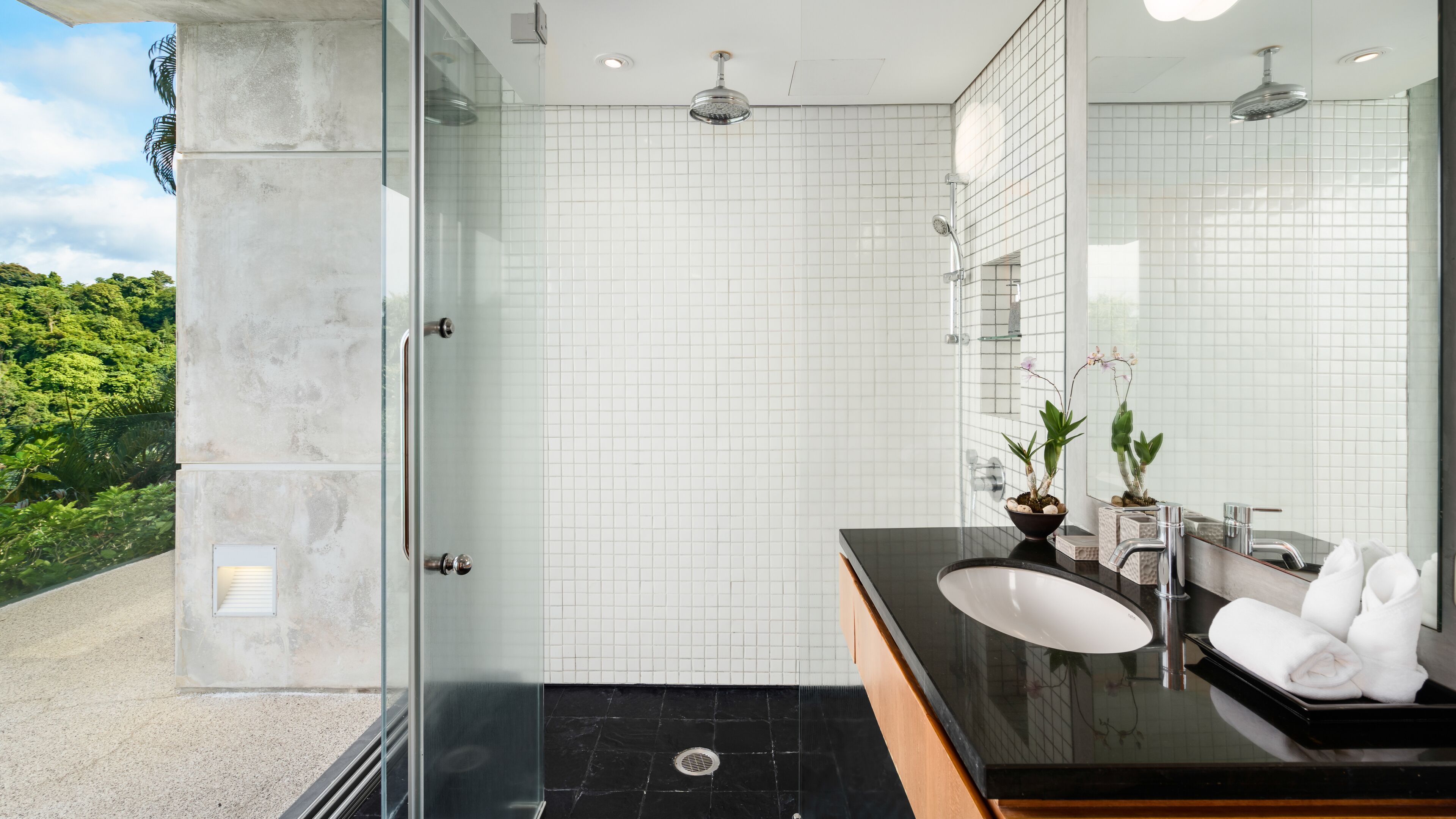 A modern bathroom with a glass shower enclosure, white tiled walls, a black countertop, and a large mirror.