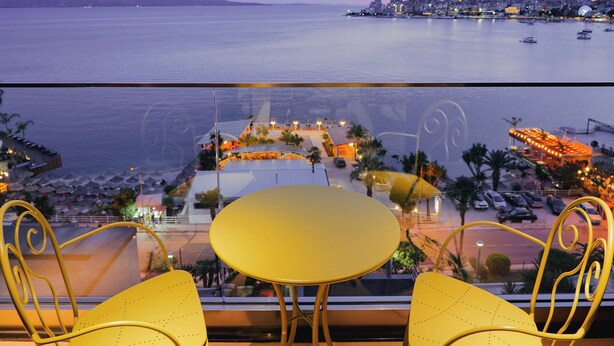 A balcony with yellow chairs and a small table, overlooking a coastal cityscape with buildings and boats.