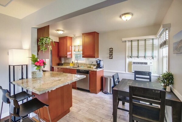 A modern kitchen with wooden cabinets, a granite countertop, and a dining area with a table and chairs. - New Bathroom! 2bd-shared yard w/spa, firepit, pets (San Diego)