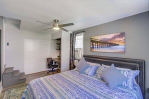 Un dormitorio con cama, escritorio, silla, ventilador de techo, ventana con cortinas y un cuadro en la pared.