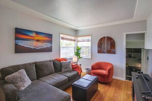 Uma sala de estar com um sofá cinzento, uma poltrona vermelha, um banquinho preto e um quadro de um pôr do sol.