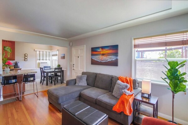 A modern living room with a gray sectional sofa, a dining area with a white table and chairs, and a large window with blinds. - New Bathroom! 2bd-shared yard w/spa, firepit, pets (San Diego)