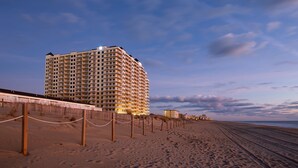 Un hotel frente a la playa con varios pisos, una playa arenosa al frente y un cielo despejado.
