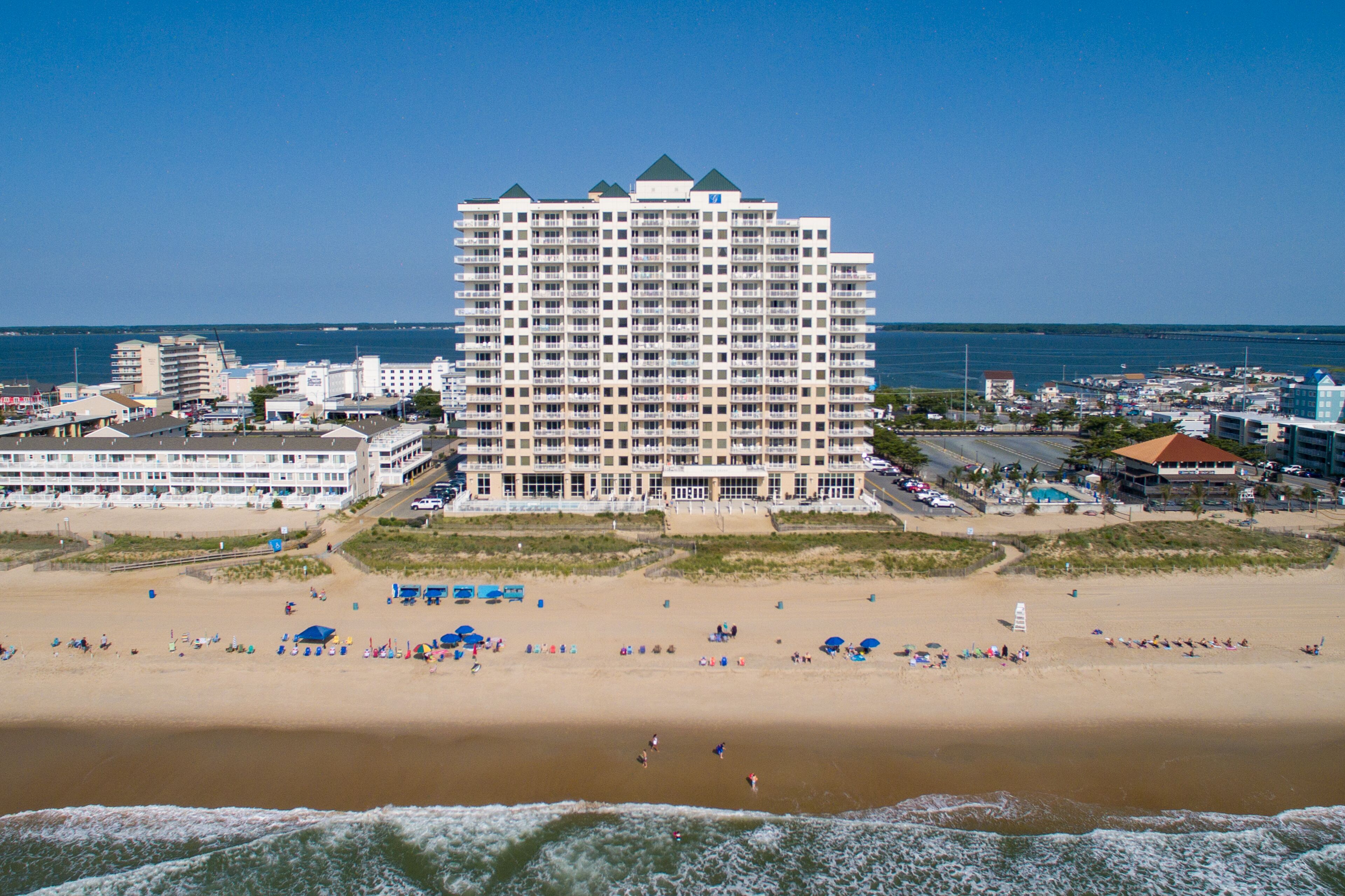 Un hotel frente a la playa con varios pisos, una playa arenosa en primer plano y vista al océano.