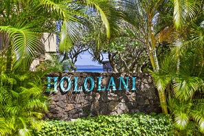 En stenvæg med navnet 'HOLOLANI' i store bogstaver, omgivet af frodig, tropisk vegetation.