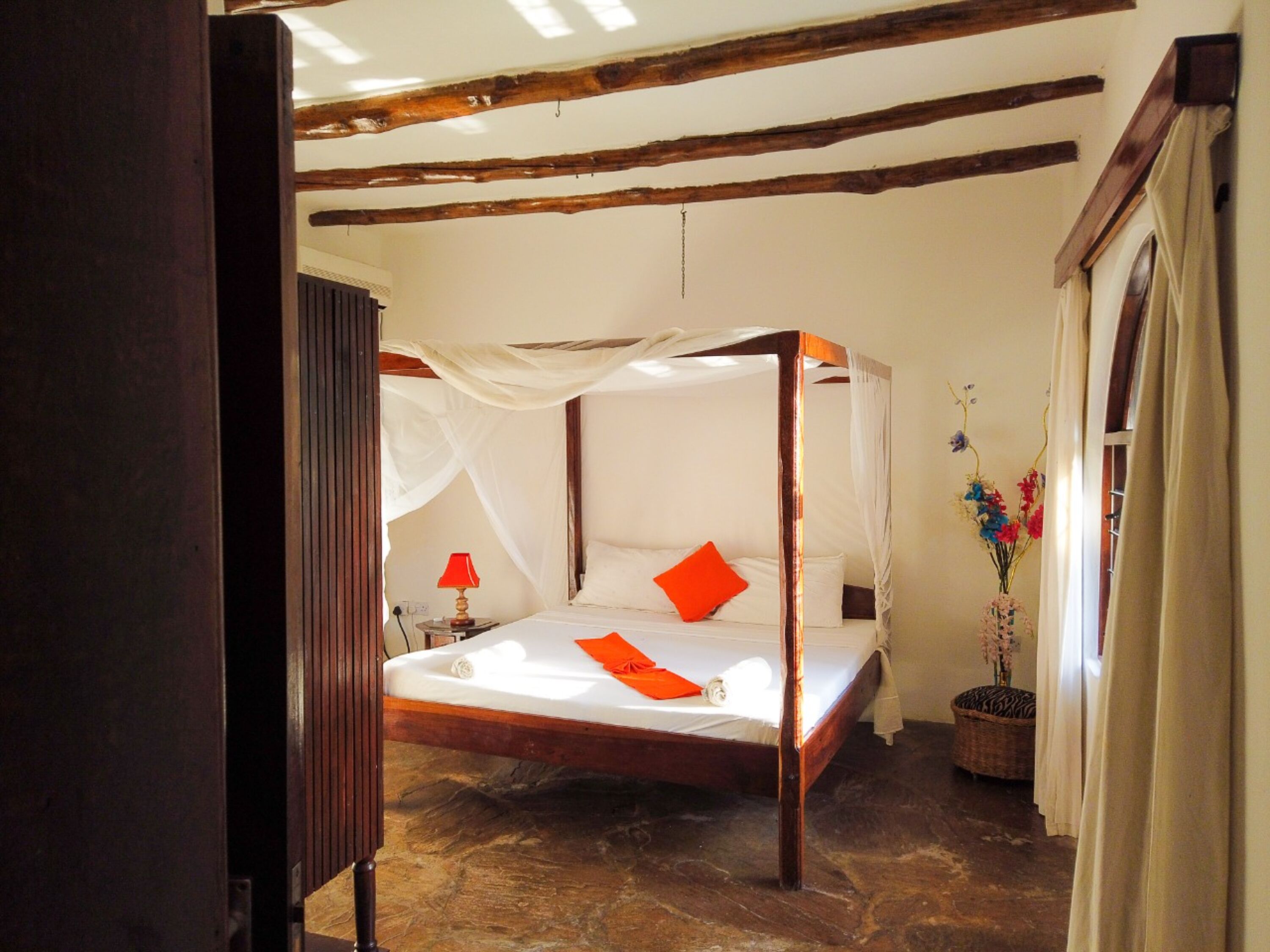 A four-poster bed with white linens and orange pillows, a red lamp on a bedside table, and a vase with flowers on the floor.
