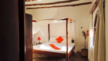 A four-poster bed with white linens and orange pillows, a red lamp on a bedside table, and a vase with flowers on the floor.