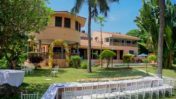 An outdoor dining area with white chairs and tables, surrounded by palm trees and a building with a balcony.