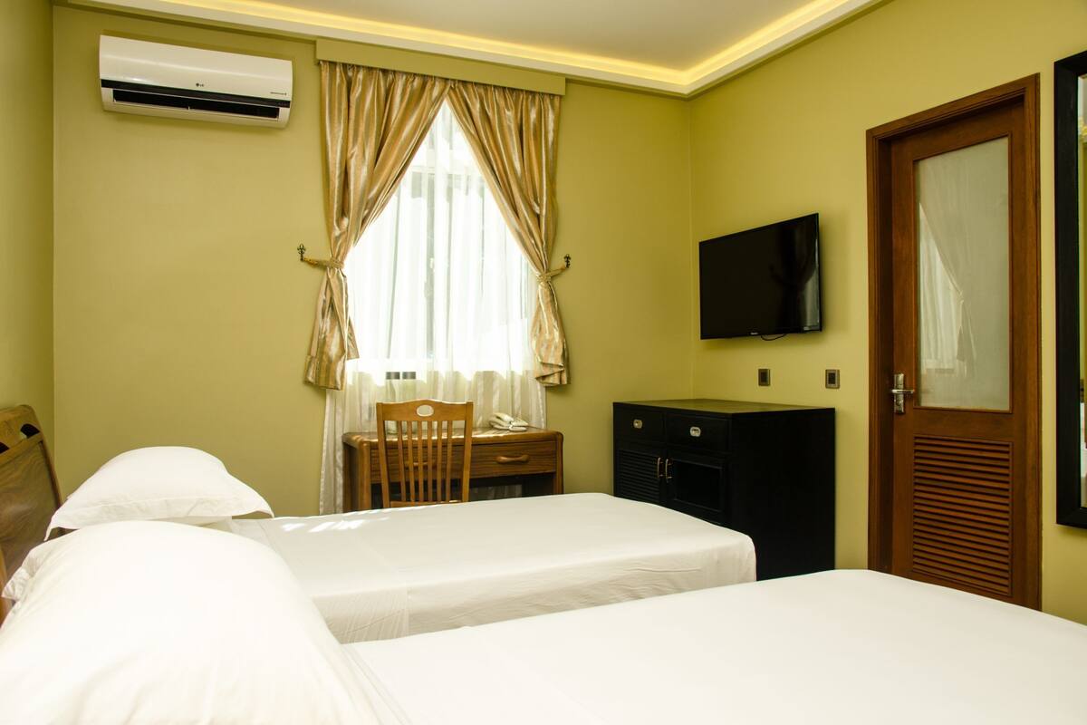 Ein Hotelzimmer mit zwei Betten, einem Schreibtisch mit Stuhl, einem an der Wand montierten Fernseher und einer Klimaanlage.